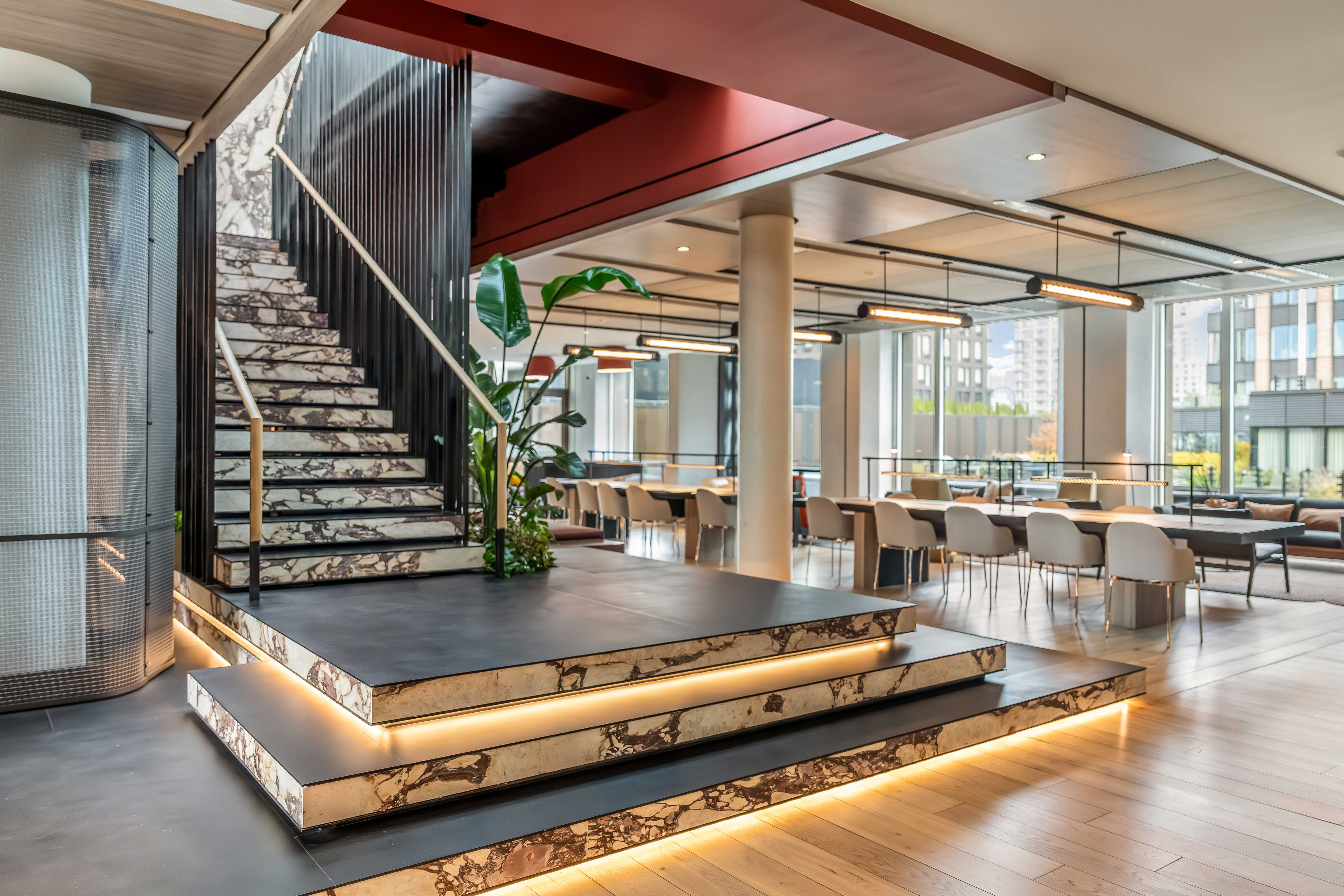 Shared lounge with an open staircase, illuminated lower steps, tables and tall windows at The Suffolk in New York City.